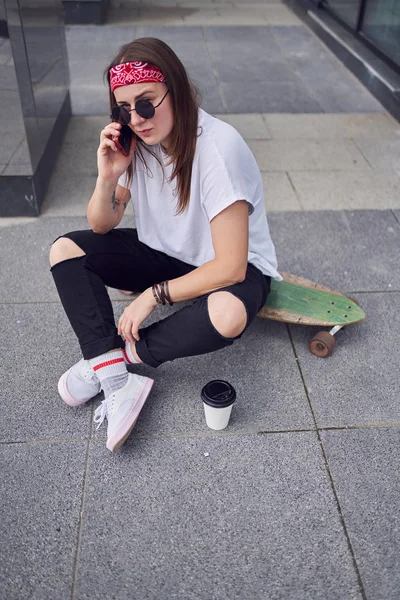 Foto de jovem morena em óculos de sol falando no telefone sentado no skate no fundo de edifícios modernos na cidade — Fotografia de Stock