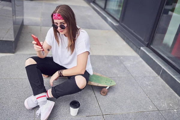 Foto di giovane bruna in occhiali da sole con il telefono in mano seduta sullo skateboard sullo sfondo di edifici moderni in città — Foto Stock