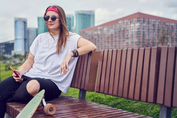 Imagem de mulher skatista com telefones em suas mãos sentado em banco de madeira contra o pano de fundo de edifícios modernos — Fotografia de Stock