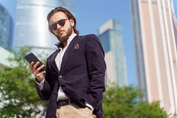 Foto de hombre en gafas de sol con teléfono en la mano mirando a la cámara para caminar en la ciudad — Foto de Stock
