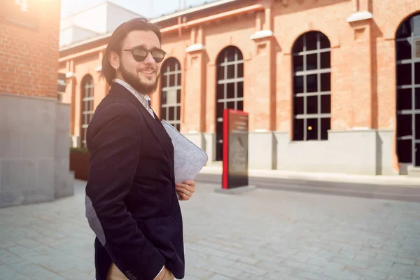 Foto de morena en gafas de sol y chaqueta con portátil de pie en la ciudad en primavera — Foto de Stock