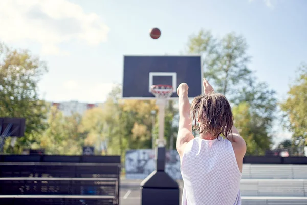 Изображение со спины спортсмена, бросающего мяч в баскетбольное кольцо на фоне голубого неба — стоковое фото