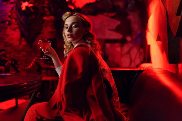 Foto de la joven rubia en chaqueta roja mirando a la cámara con cóctel en la mano en el club nocturno — Foto de Stock
