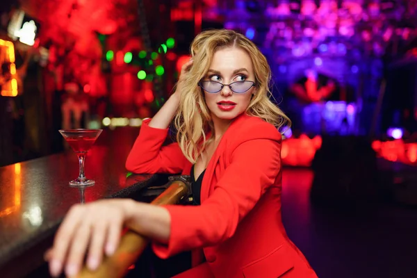 Foto de la joven rubia con gafas y traje rojo mirando al lado sentado en el bar con copa de cóctel en la discoteca — Foto de Stock