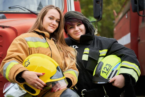 Foto de duas mulheres bombeiras com capacete em suas mãos ao lado do carro de bombeiros — Fotografia de Stock