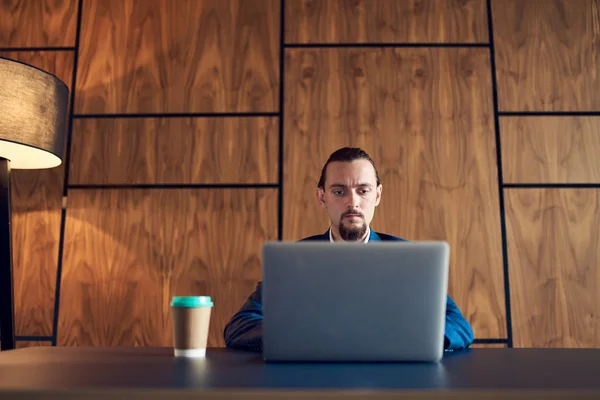 Obrázek seriózního muže sedícího u stolu s notebookem a papírovým kelímkem s kávou na pozadí dřevěné stěny . — Stock fotografie