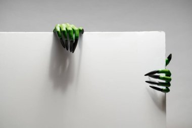 Uzun tırnaklı iki yeşil zombi eli, cadılar bayramı için stüdyoda gri arka planda boş bir kağıt tutuyorlar..