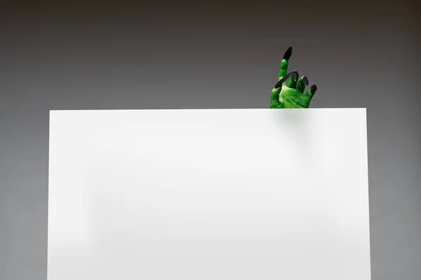 Mano zombi verde con clavos largos sostiene hoja de papel en blanco sobre fondo gris en estudio —  Fotos de Stock