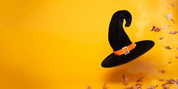 Black witch hat, dry autumn leaves on blank orange background, space for text. — 图库照片