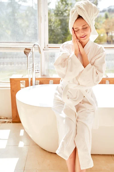 Bild einer Frau mit geschlossenen Augen im weißen Bademantel und mit Handtuch auf dem Kopf, die in der Nähe des Bades in einem Raum mit großem Fenster steht — Stockfoto