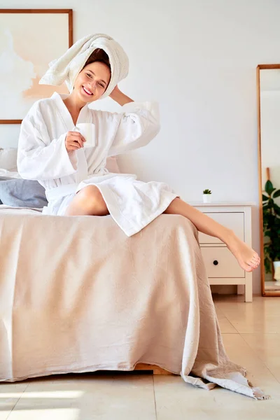 Zdjęcie uśmiechniętej kobiety w białym szlafroku z kubkiem kawy w rękach leżącej na łóżku . — Zdjęcie stockowe