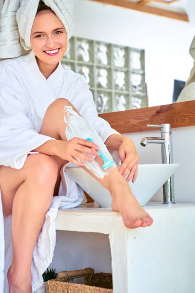 Ganzkörperbild einer glücklichen Frau im weißen Mantel, die sich im Bad die Beine rasiert — Stockfoto