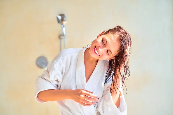 Imagem de menina morena sorridente com cabelo molhado em roupão de banho branco em pé na banheira . — Fotografia de Stock