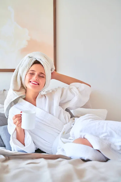 Zdjęcie szczęśliwej kobiety w białym szlafroku z kubkiem herbaty w rękach leżącej na łóżku. — Zdjęcie stockowe