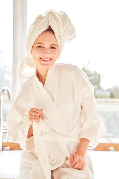 Portrait of happy girl in white bathrobe and with towel on her head standing near bath in room with large window — Stock Photo, Image