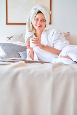 Beyaz bornozlu, elinde bir kupa çayla yatağa uzanmış gülümseyen bir kadın fotoğrafı..