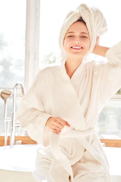 Image of happy girl in white bathrobe and with towel on her head standing near bath in room with large window — Stock Photo, Image