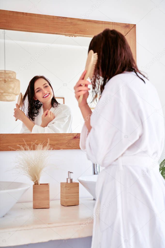 Image of brunette combing her hair in front of bathtub mirror