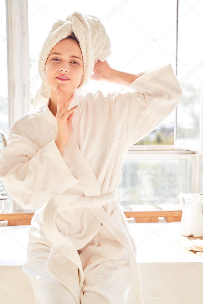 Image of girl in white bathrobe and with towel on her head standing near bath in room with large window in afternoon