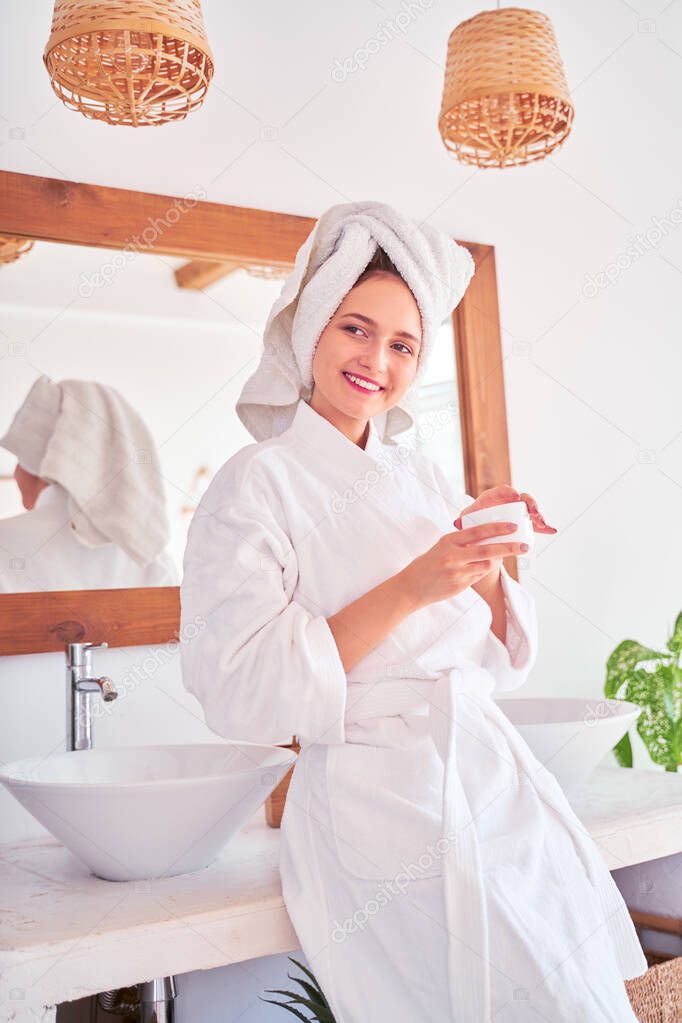 Photo of young woman in bathrobe with cream in her hands in bathroom.