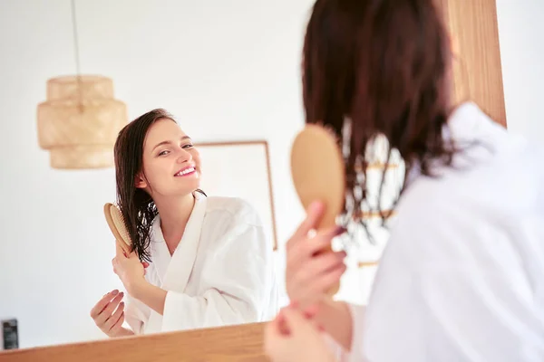 Photo of brunette combing her hair in front of bathtub mirror — Stock Photo, Image