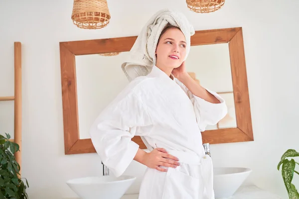 Obraz młodej kobiety w białym szlafroku z ręcznikiem na głowie stojącej w łazience na tle lustra — Zdjęcie stockowe