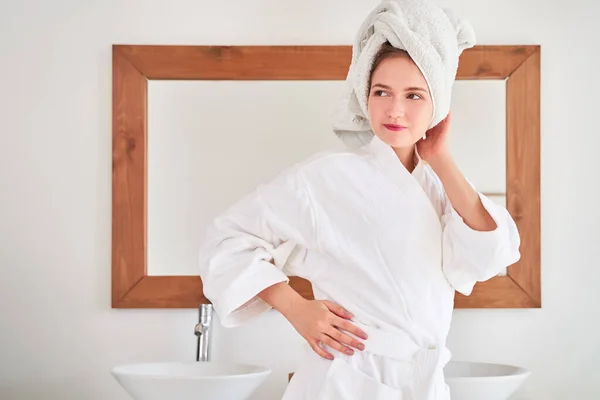 Zdjęcie młodej kobiety w białym szlafroku z ręcznikiem na głowie stojącej w łazience na tle lustra — Zdjęcie stockowe