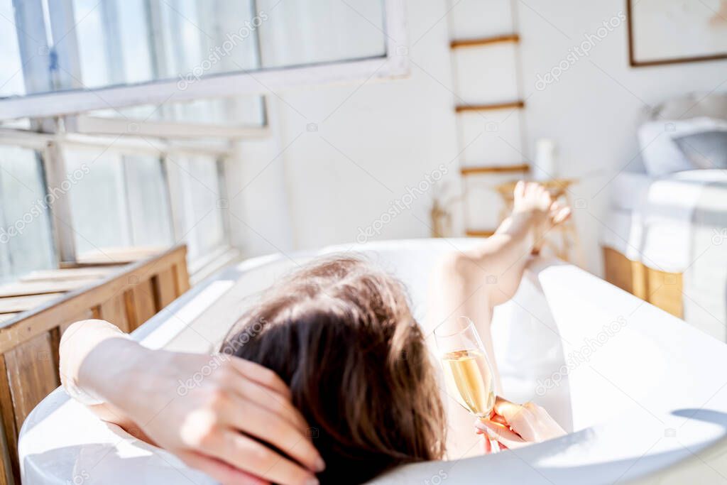 Image from back of young woman with wine glass with wine in her hands lying in bath.