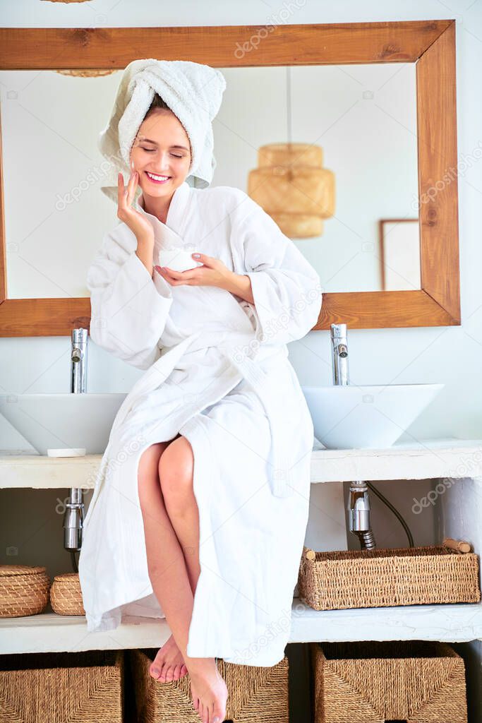 Photo of young woman with bathrobe with cream in her hands standing in bathroom.