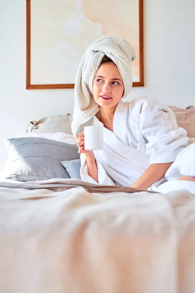 Zdjęcie kobiety w białym szlafroku z kubkiem herbaty w rękach leżącej na łóżku. — Zdjęcie stockowe