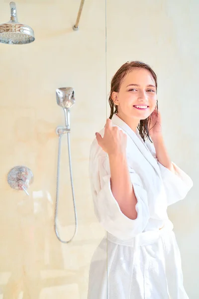 Foto de mulher morena feliz com cabelo molhado em roupão de banho branco olhando para a câmera enquanto estava de pé no banho . — Fotografia de Stock