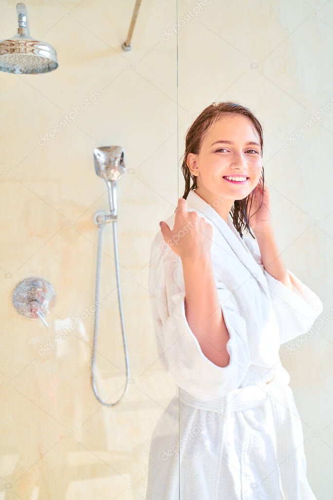 Photo of happy brunette woman with wet hair in white bathrobe looking at camera while standing in bath.