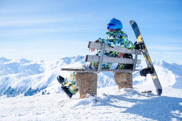 Foto de atleta sentado en el banco con snowboard en el resort de invierno . —  Fotos de Stock