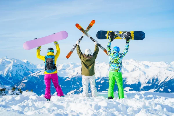 Zdjęcie z tyłu trzech sportowców z ramionami podniesionymi snowboardami i nartami w dzień . — Zdjęcie stockowe