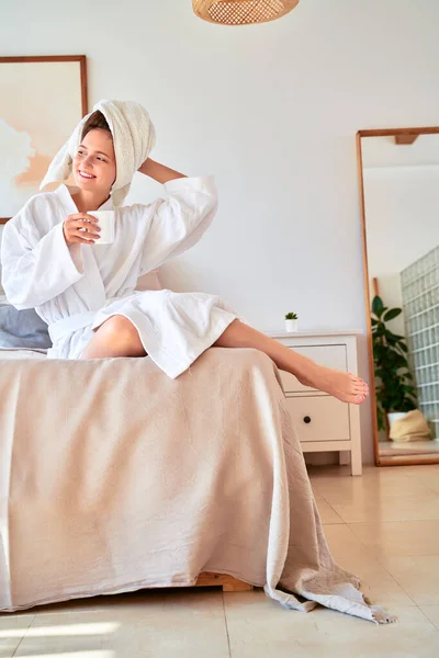 Obraz uśmiechniętej dziewczyny w białym szlafroku z kubkiem kawy w rękach leżącej na łóżku. — Zdjęcie stockowe