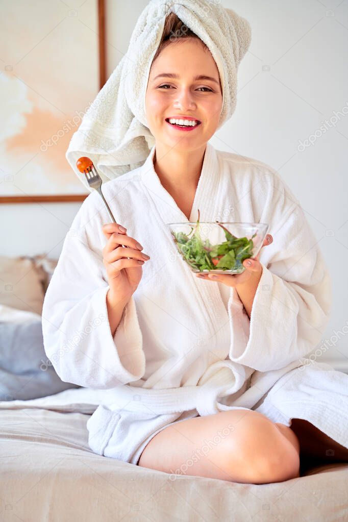 Image of young girl in bathrobe with salad in bowl in her hands sitting on bed.