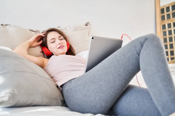 Immagine di una giovane donna bruna con tablet in mano che ascolta musica mentre è sdraiata sul letto . — Foto Stock