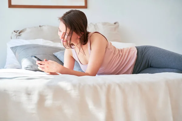 Изображение юной брюнетки с телефоном в руках, лежащей на кровати — стоковое фото