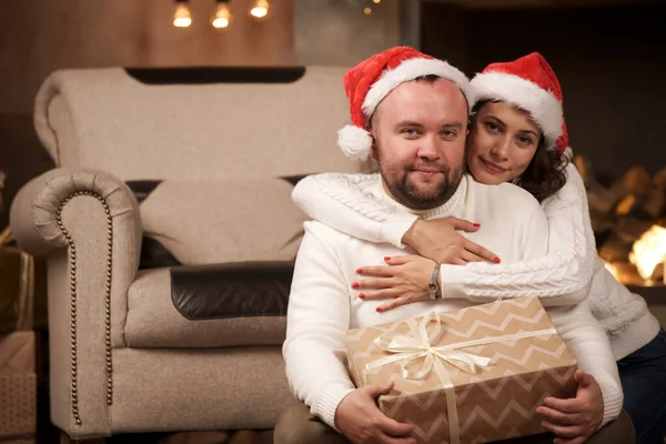 Foto de hombres y mujeres abrazados en gorras de Santas sentados en el suelo en la habitación con el interior de Navidad — Foto de Stock