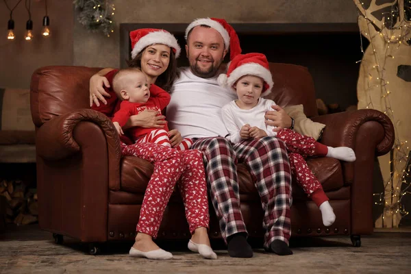 Zdjęcie szczęśliwych rodziców w czapkach Mikołaja i synów na skórzanej sofie w studio — Zdjęcie stockowe
