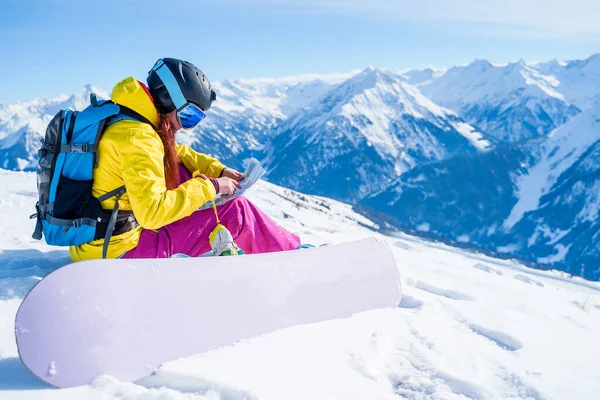 Foto lateral de una joven deportista con mapa en sus manos y tabla de snowboard sentada en la ladera nevada . —  Fotos de Stock