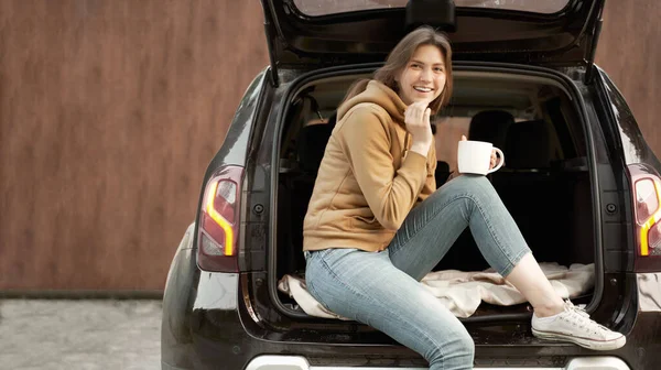 Morena feliz con taza sentada en el maletero abierto de coche negro en el día de verano —  Fotos de Stock