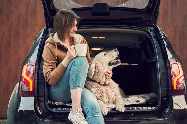 Ragazza felice con tazza in mano con cane seduto nel bagagliaio aperto della macchina nera. — Foto Stock