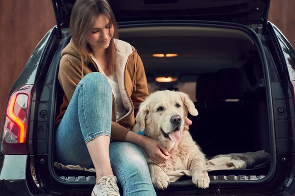 Mujer joven con golden retriever sentarse en el maletero abierto de coche negro — Foto de Stock