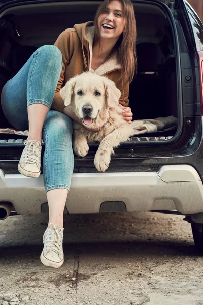 快乐的女人拥抱着金发碧眼的猎犬，坐在开着的黑色轿车后备箱里 — 图库照片