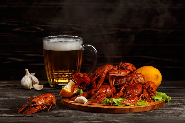 Sıcak haşlanmış kerevit onlardan gelen Buhar ile bir tabak kahverengi ahşap arka plan üzerinde bir bira bardağı yanında duruyor. Çerçevede orada da limon ve sarımsak.