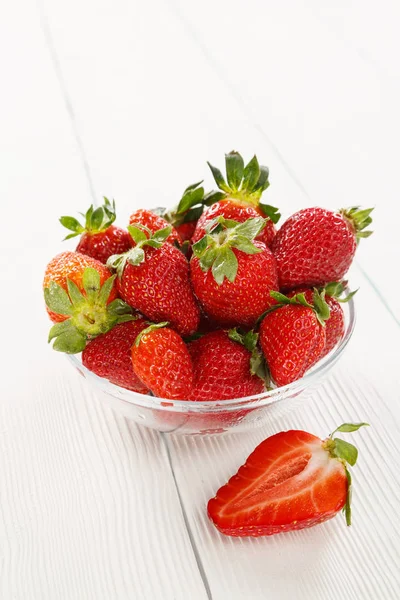 一个成熟的花园草莓的照片 在一个玻璃碗 它站在一个纹理的白色木桌顶部 明亮的夏日阳光 一个切浆果躺在盘子旁边 图库照片