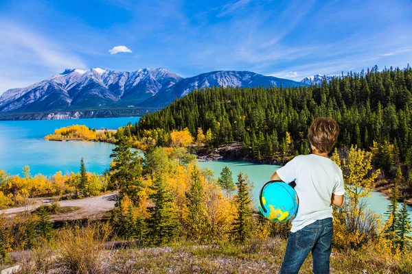 Exquisite Abraham Lake with turquoise water. Nine-year-old boy in jeans with a globe in his hands admires the lake. Indian Summer in the Rockies of Canada. Concept of ecological and active tourism