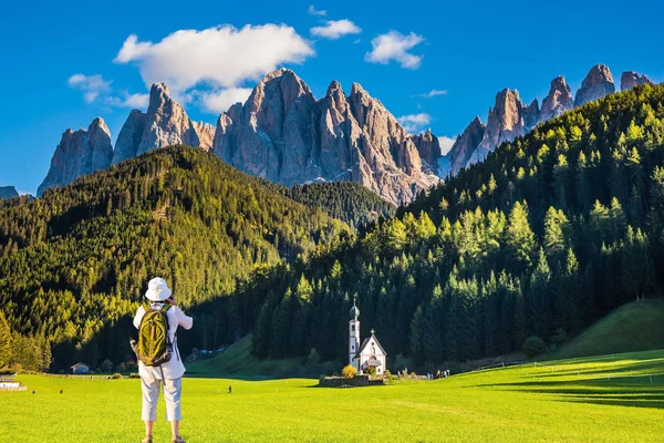 Dolomites, Tirol. Active woman-tourist with backpack photographs the church of Santa Maddalena. Forested mountains surrounded by green Alpine meadows. The concept of ecological and active tourism