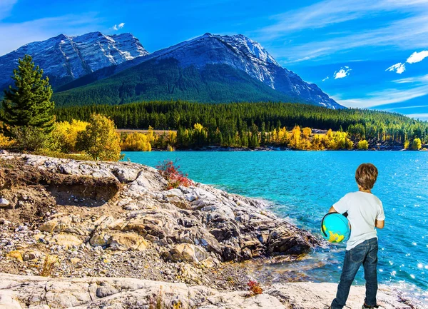 Exquisite Abraham Lake with turquoise water. Nine-year-old boy in jeans with a globe in his hands admires the lake. Indian Summer in the Rockies. Concept of ecological and active tourism